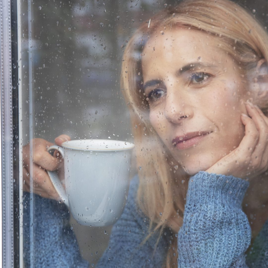 mature-woman-looking-outside-her-window-while-it-rains-drinking-tea